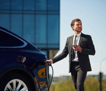 Holding the charger. Businessman is standing near his electric car outdoors